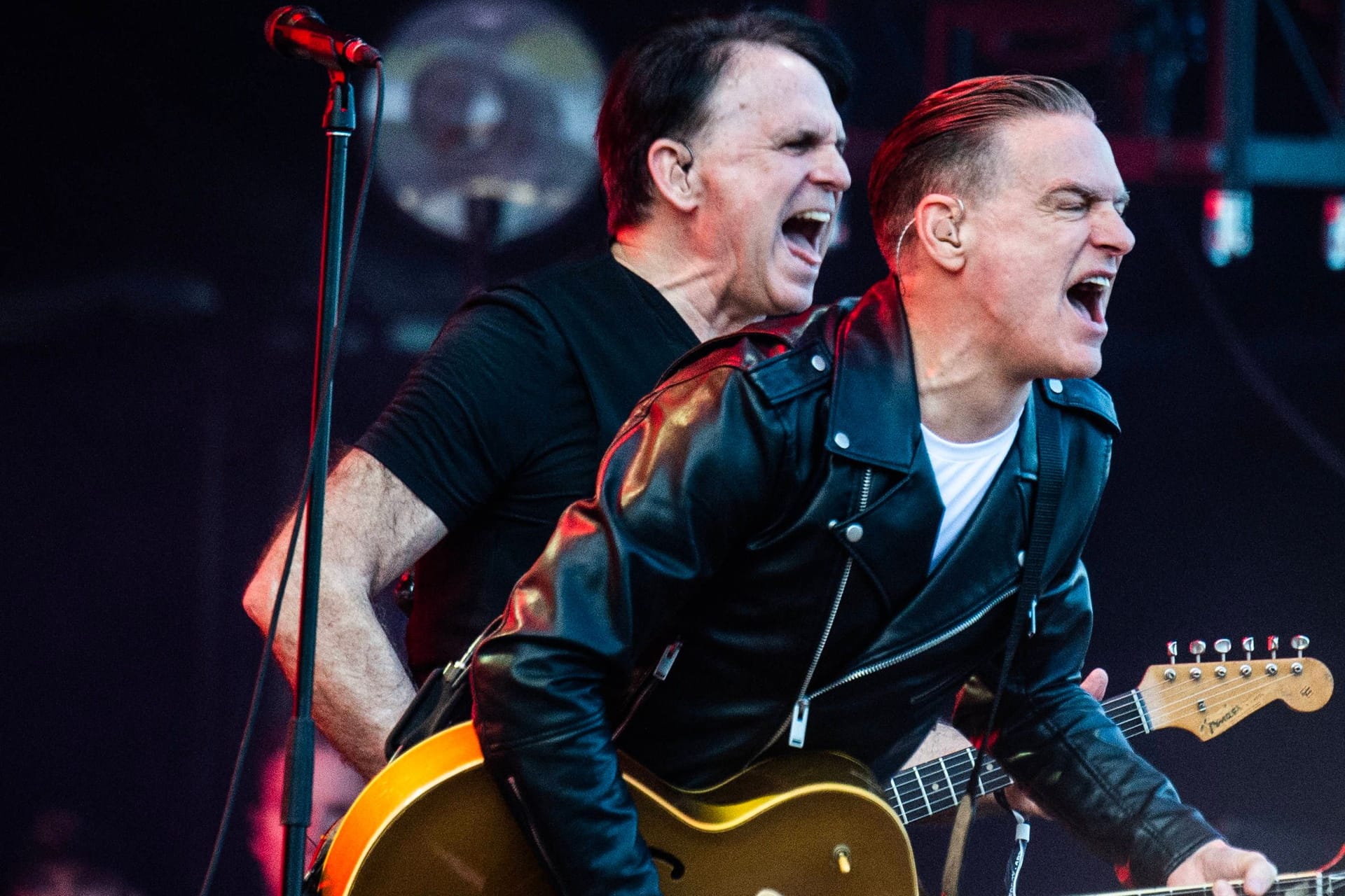 Bryan Adams (r.) und Keith Scott stehen auf einer Bühne. Beide Musiker spielen auf einer Gitarre und singen. Bryan Adams trägt eine schwarze Lederjacke, Keith Scott trägt ein schwarzes T-Shirt. Links und rechts von ihnen steht jeweils ein Mikrofon. Im Hintergrund sind Scheinwerfer zu erkennen.