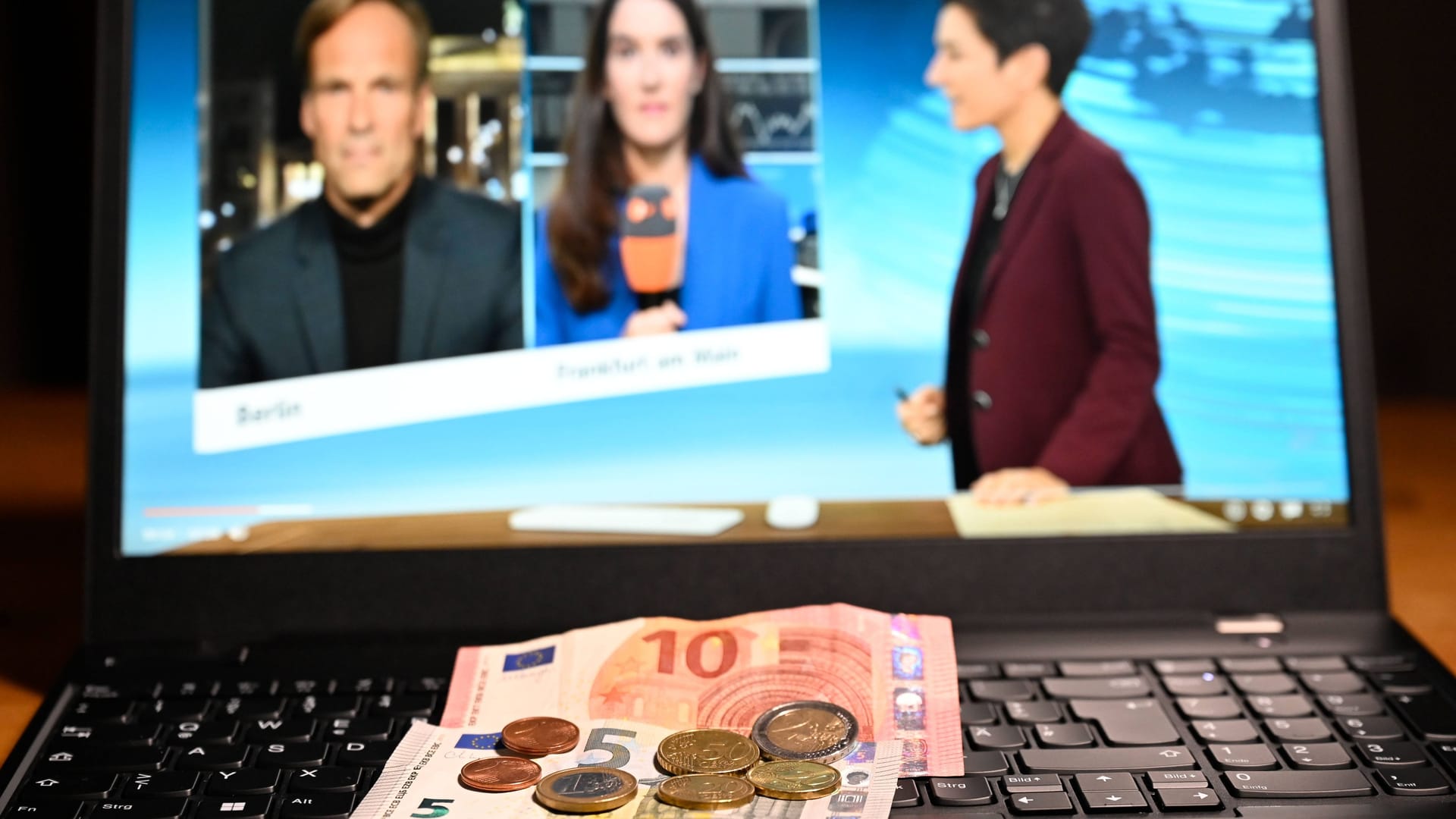 Bargeld liegt auf einem Laptop. Auf dem Bildschirm ist ein Ausschnitt des ZDF-Heute-Journals zu sehen, Moderatorin ist Dunja Hayali.