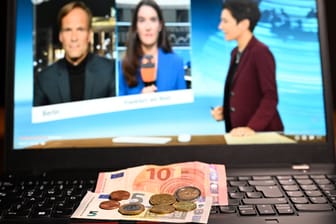 Bargeld liegt auf einem Laptop. Auf dem Bildschirm ist ein Ausschnitt des ZDF-Heute-Journals zu sehen, Moderatorin ist Dunja Hayali.