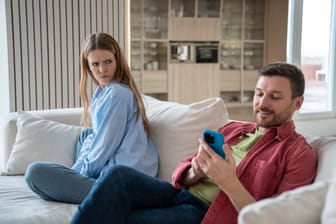 Wife angry at husband who looking at smartphone screen smiling sitting on couch at home.