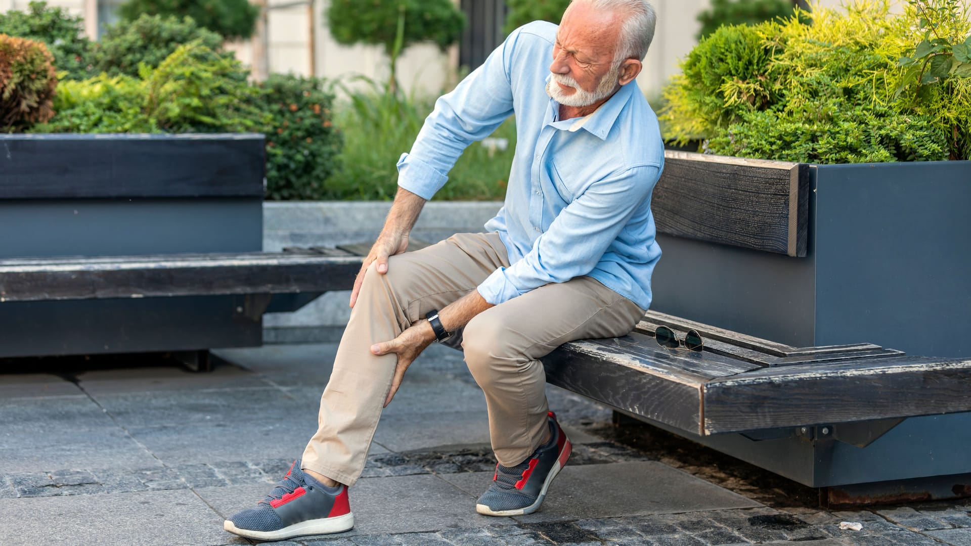 Ein älterer Mann sitzt auf einer Bank und hält sich Knie und Wade.