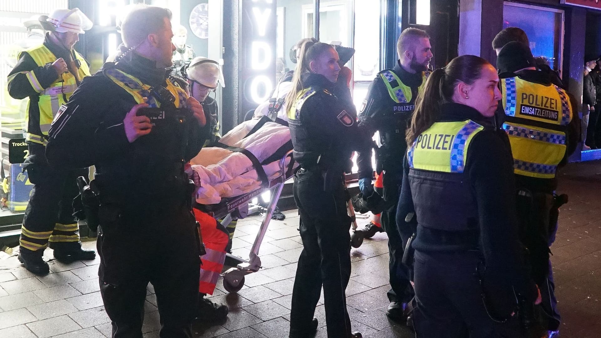 Einsatzkräfte der Polizei stehen neben einem auf einer Trage liegenden Verletzten vor einem Friseursalon in Hamburg-Billstedt.