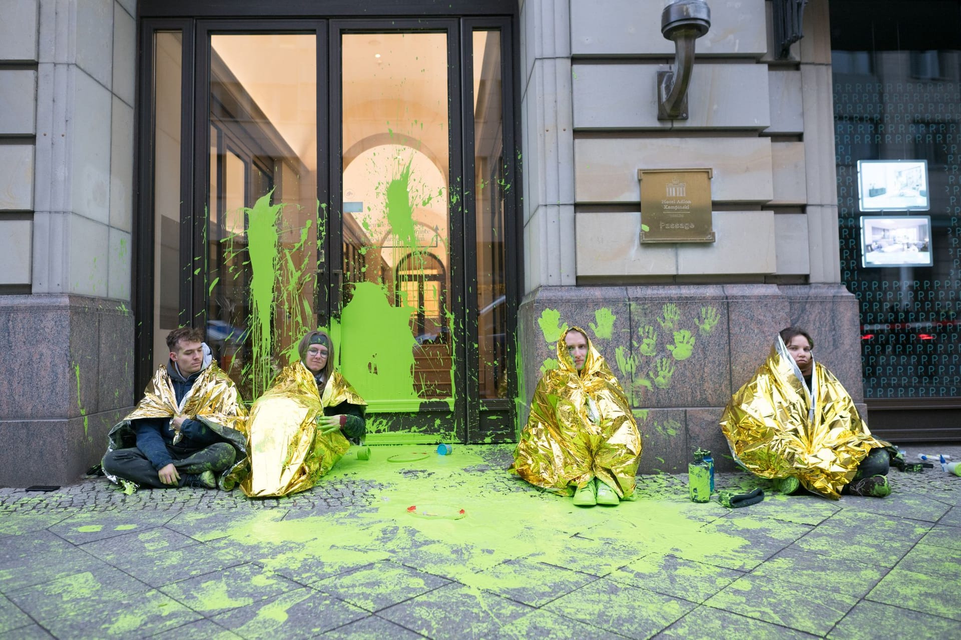 10.12.2024, Berlin: Aktivisten der Letzten Generation sitzen während einer Demonstrationen gegen den "World LNG Summit" der Gasindustrie und Flüssiggasanbieter in Berlin vor einem Eingang des Adlon, der mit grüner Farbe beschmiert ist. Der Gasenergiegipfel findet vom 9. bis 12. Dezember im Hotel Adlon statt.