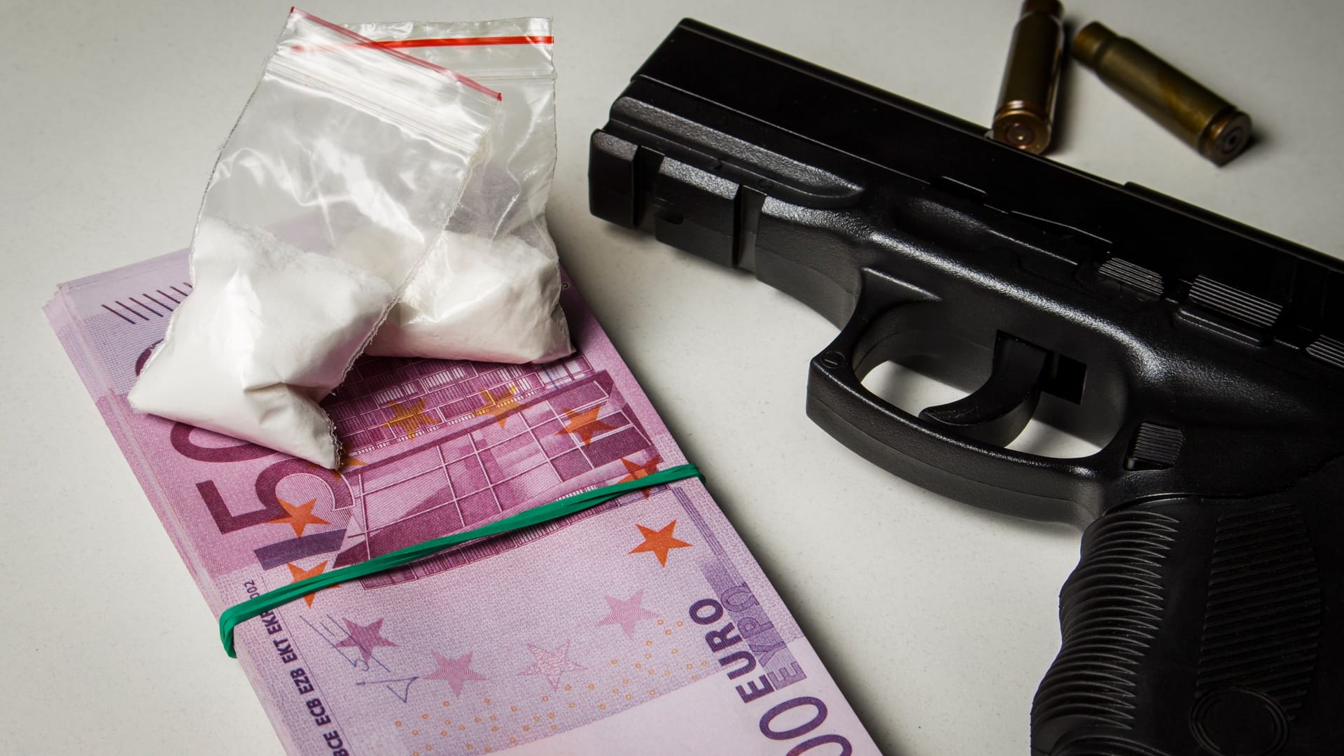 Black gun with a pack of euros and packets of drugs on a white table