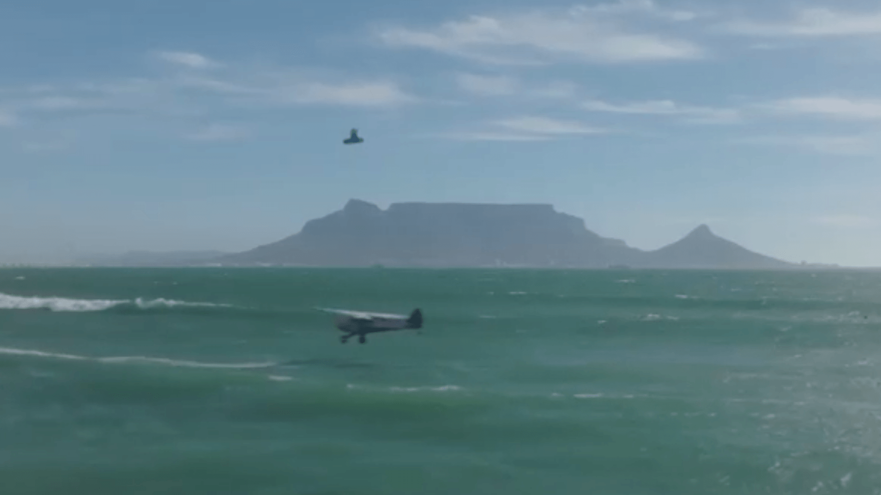 Ein Kitesurfer springt über ein Flugzeug, das ganz nah an der Wasseroberfläche fliegt.