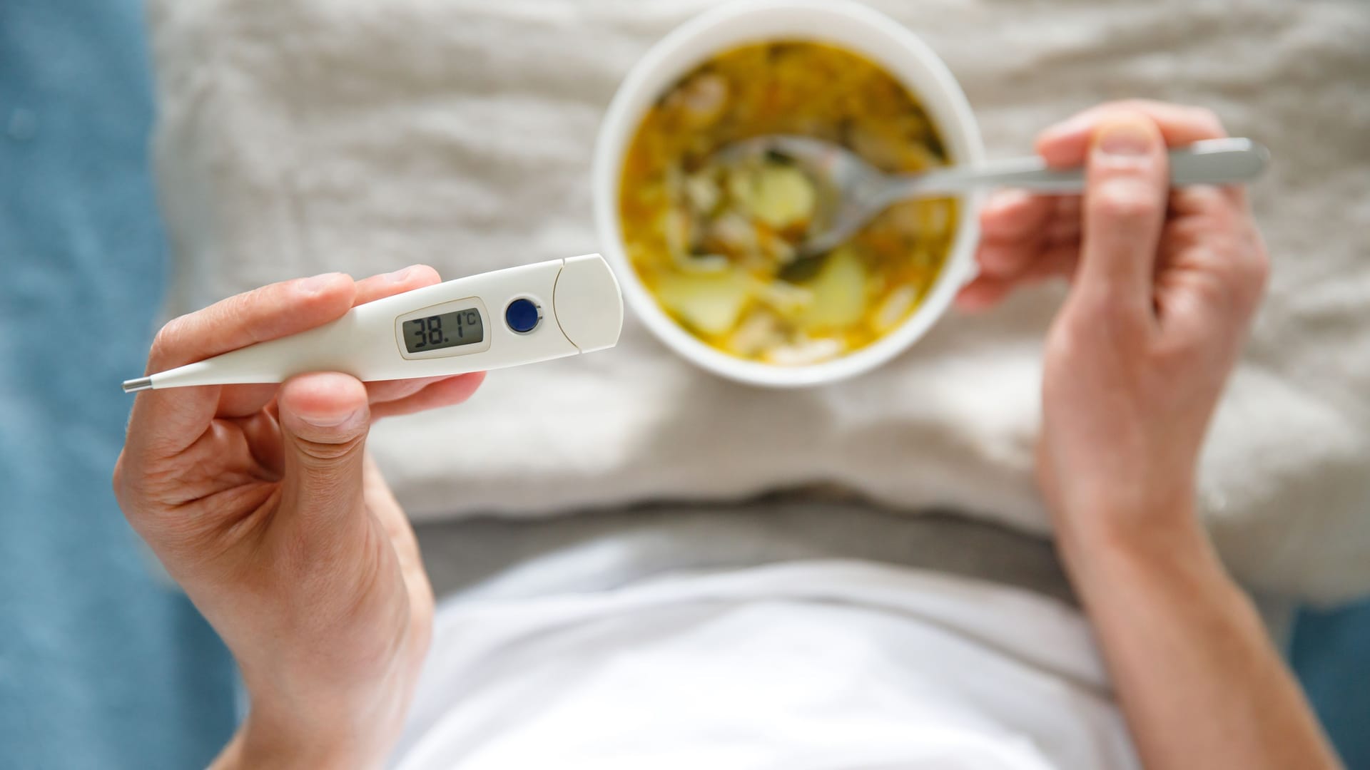 Jemand liegt mit Fieberthermometer und Suppe im Bett: Bei Fieber kommt vor allem leichte Kost infrage.