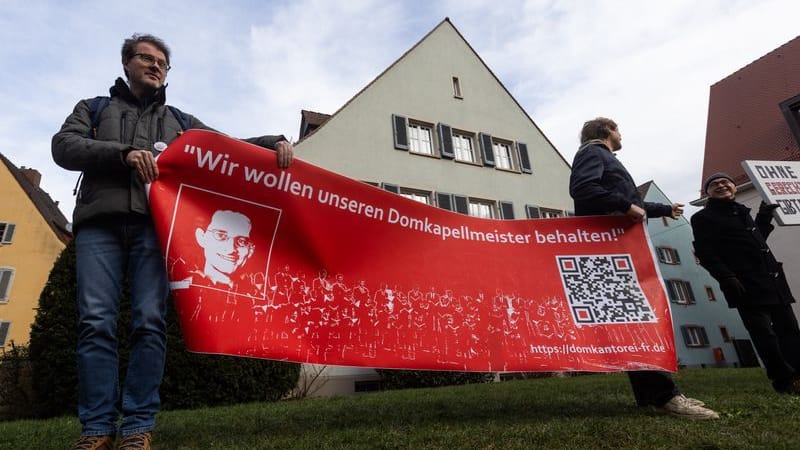 Demonstranten stehen nach einem Gottesdienst hinter dem Freiburger Münster und warten mit einem Banner mit der Aufschrift "Wir wollen unseren Domkapellmeister behalten" auf den Erzbischof.