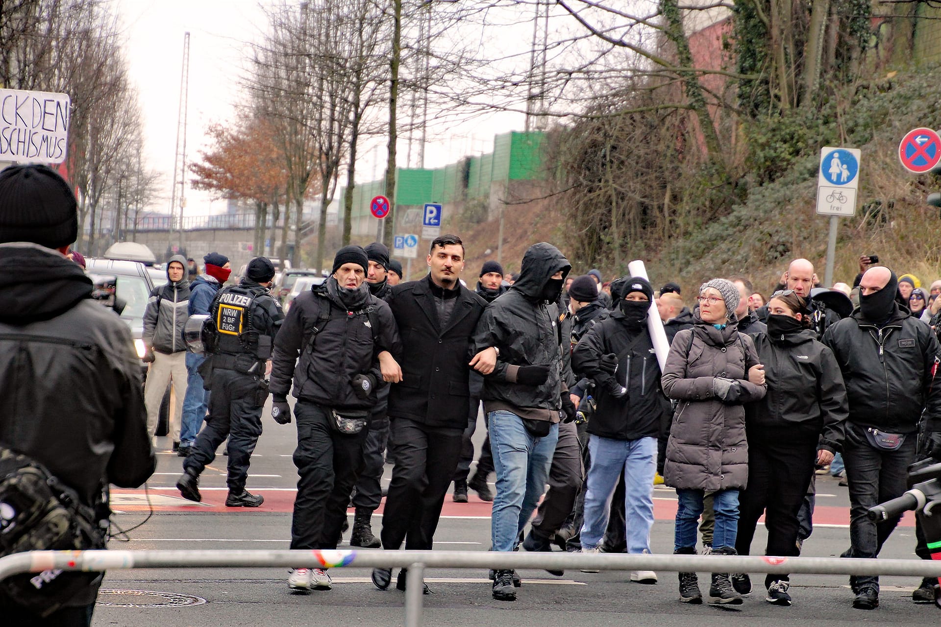 Arm in Arm eingehakt: Ex-AfD-Politiker Ferhat Sentürk (mittig, schwarzer Mantel) läuft zusammen mit Teilnehmern der Demo sowie der AfD-Funktionärin Gisela Ellgoth (rechts im Bild, grauer Mantel) durch Aachen.