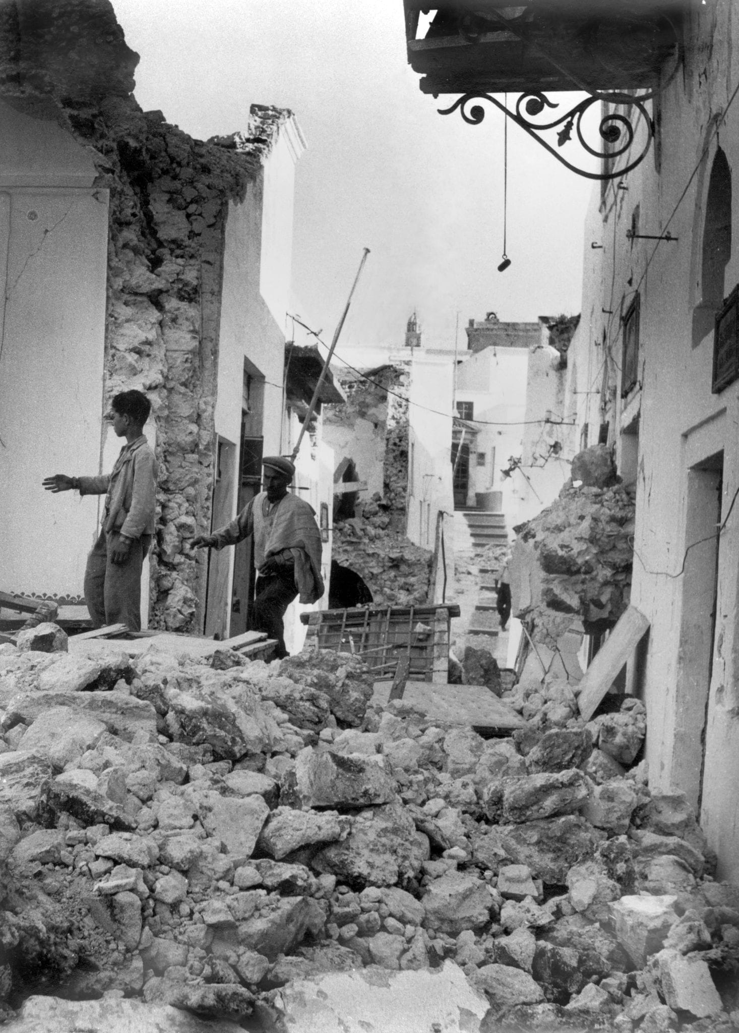 Zwei Bewohner gehen über Trümmer von Wohnhäusern durch eine kleine, völlig verwüstete Ortschaft auf der Insel Santorini am 12.07.1956