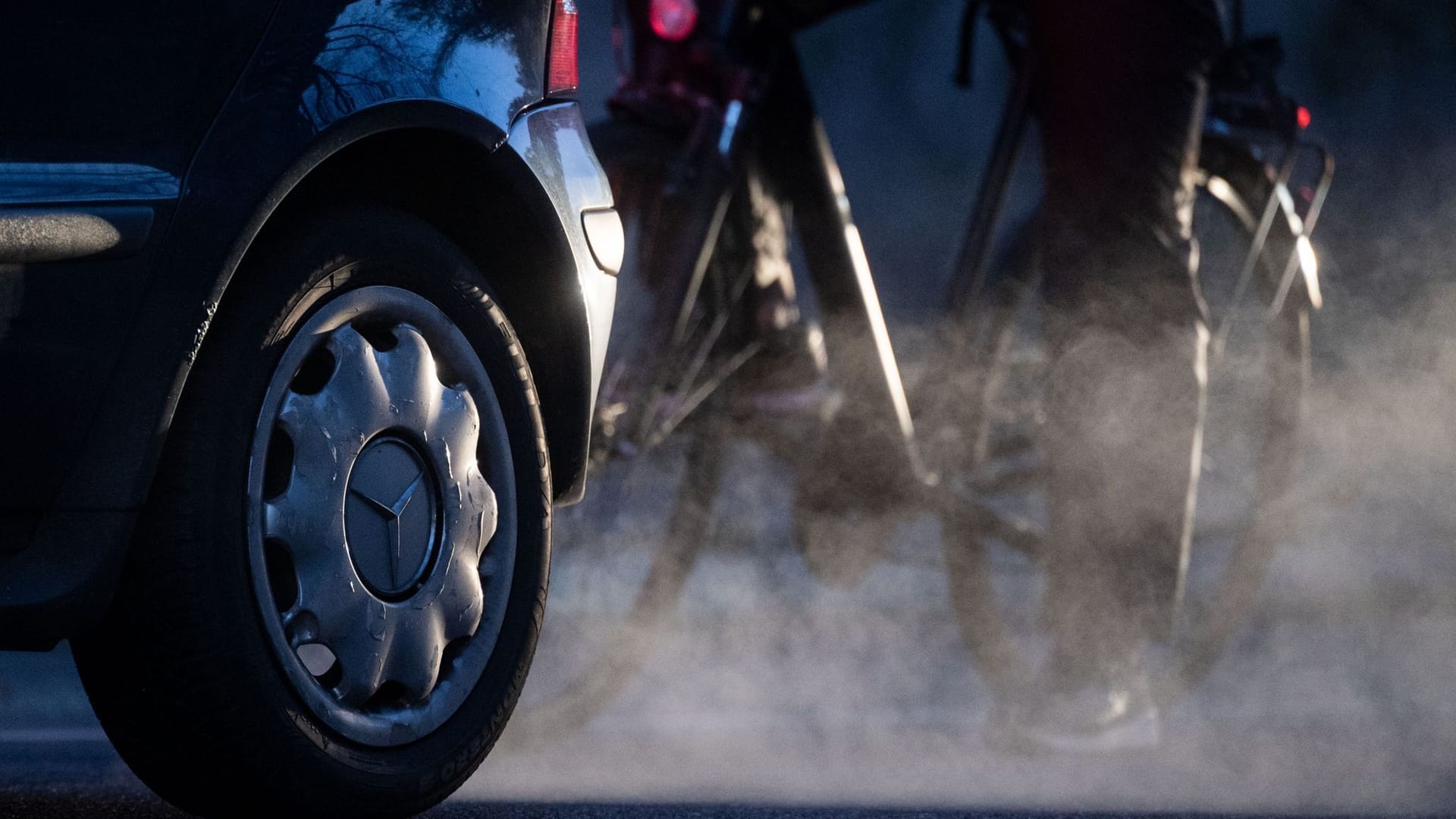 Ein Radfahrer steht neben einem Auto von Mercedes mit Dieselantrieb, dessen Abgase in der kalten Morgenluft sichtbar werden.