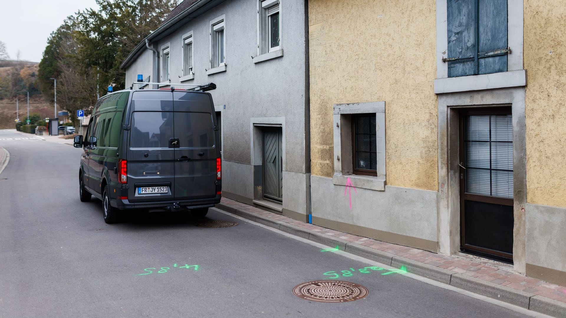 Markierungen der Kriminaltechnik sind auf einer Straße zu sehen, während ein mit Farbe gesprühter Pfeil der Kriminaltechnik auf ein Einschussloch in einem Fenster hinweist.