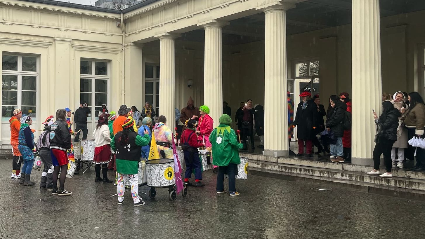 Eine Trommelgruppe lässt sich die Laune von dem plötzlich einsetzenden Regen nicht vermiesen: Unter dem Dach des Elisenbrunnens suchen sie Schutz.