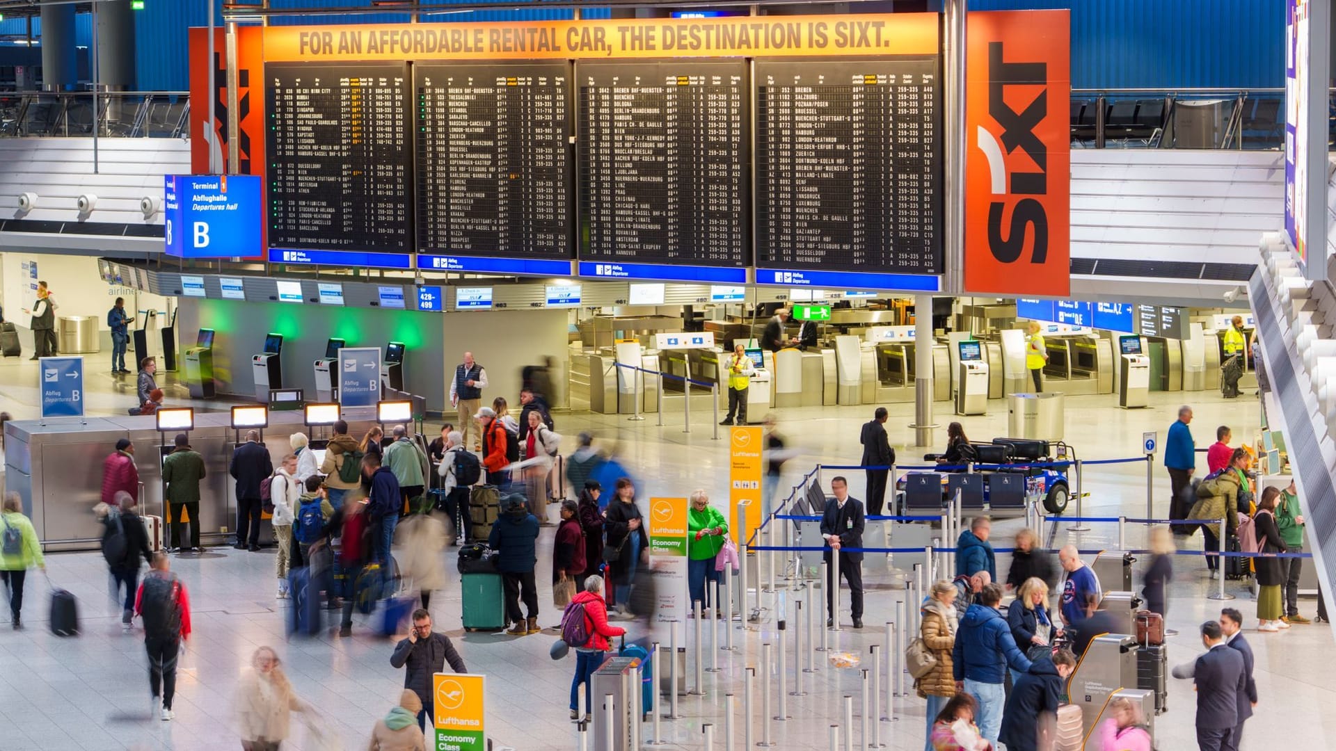 Passagiere gehen durch Terminal 1 am Flughafen Frankfurt. Im Hintergrund der Menschenmenge ist die große Anzeigetafel mit Abflügen und Ankünften zu sehen.