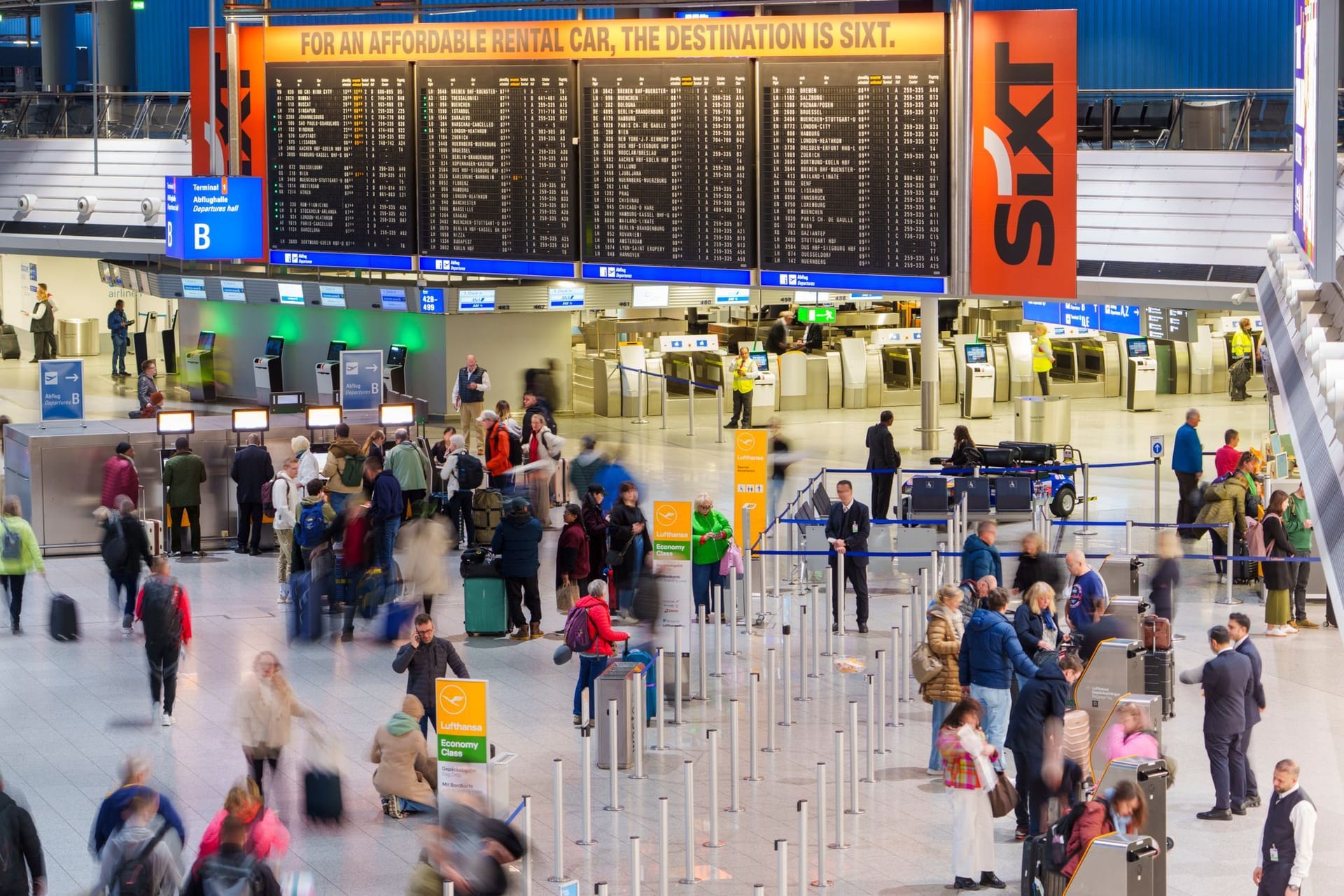 Passagiere gehen durch Terminal 1 am Flughafen Frankfurt. Im Hintergrund der Menschenmenge ist die große Anzeigetafel mit Abflügen und Ankünften zu sehen.