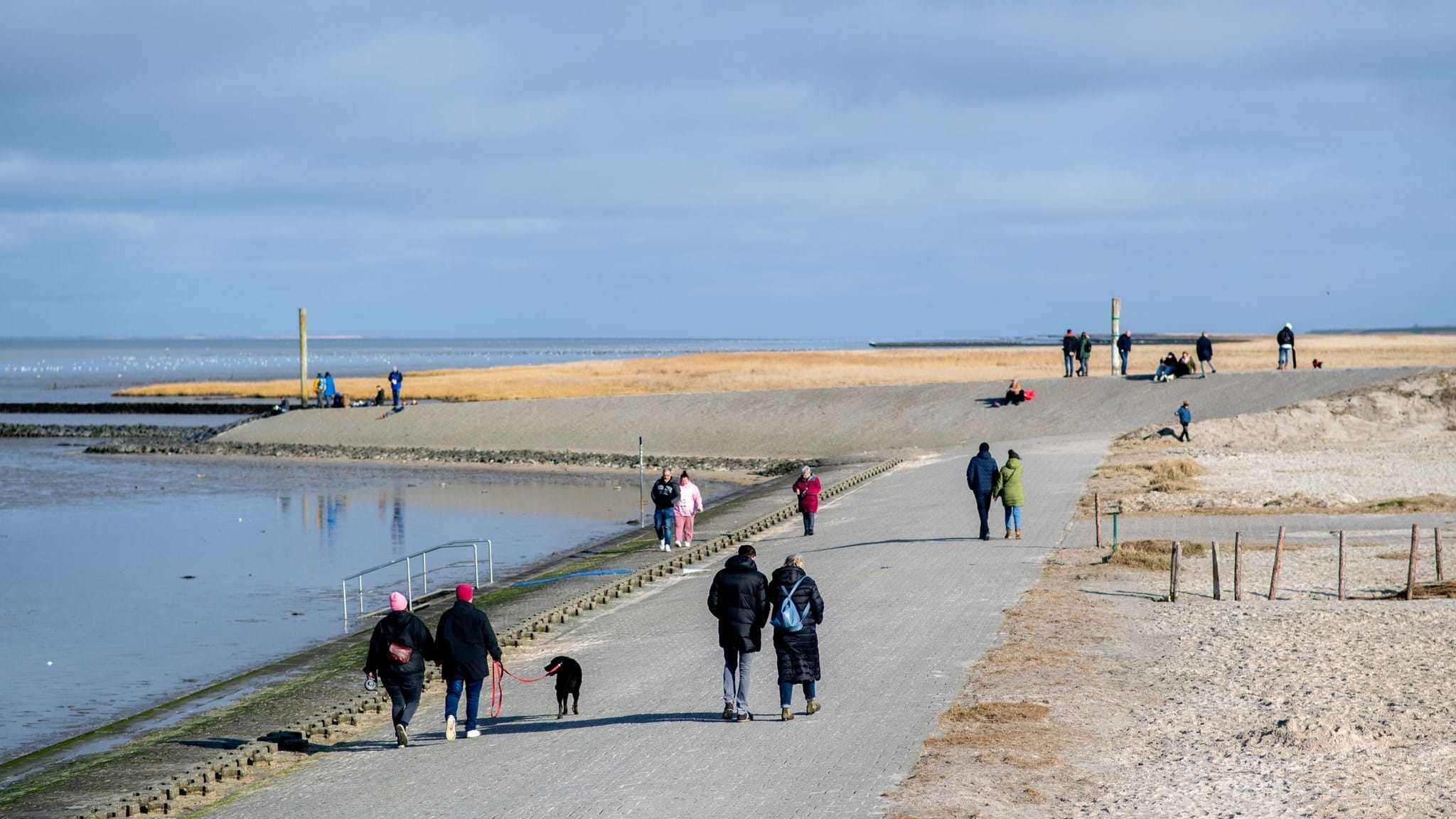 Menschen gehen bei sonnigem Wetter auf einer Mole an der Hafeneinfahrt von Harlesiel entlang. Zum meteorologischen Frühlingsbeginn wird das Wetter freundlicher.