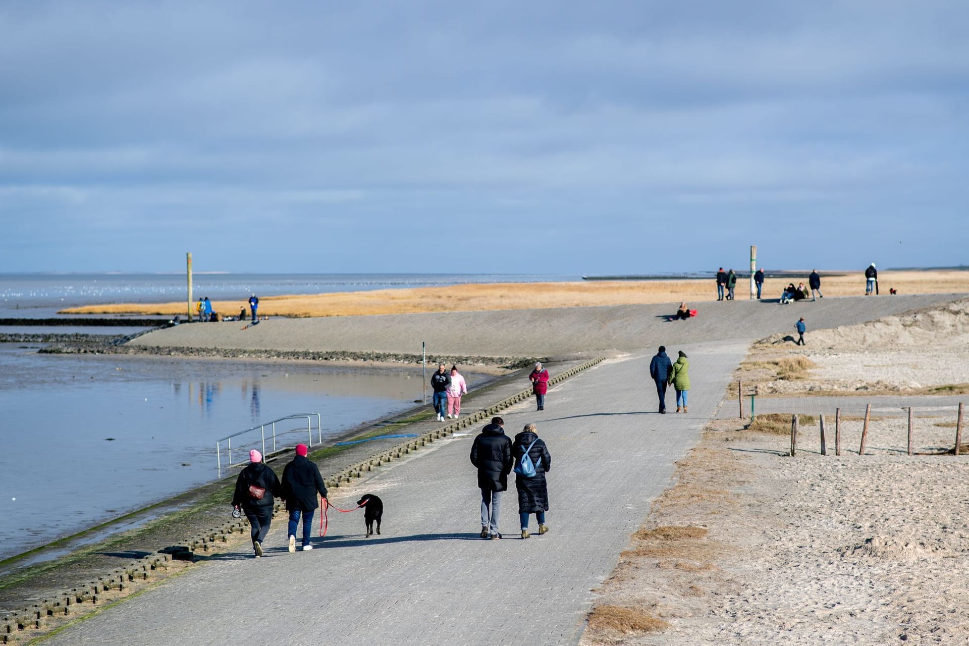 Menschen gehen bei sonnigem Wetter auf einer Mole an der Hafeneinfahrt von Harlesiel entlang. Zum meteorologischen Frühlingsbeginn wird das Wetter freundlicher.