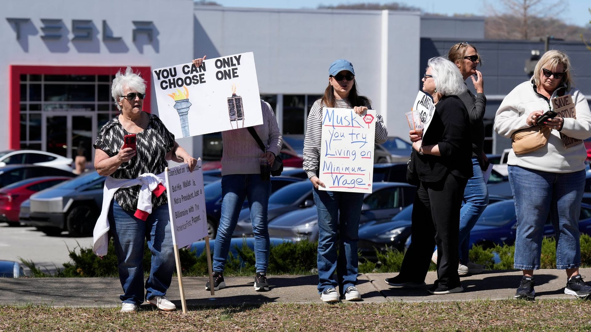 Demonstranten tragen Schilder und skandieren Slogans gegen Elon Musk und Präsident Donald Trump vor einem Tesla-Händler.