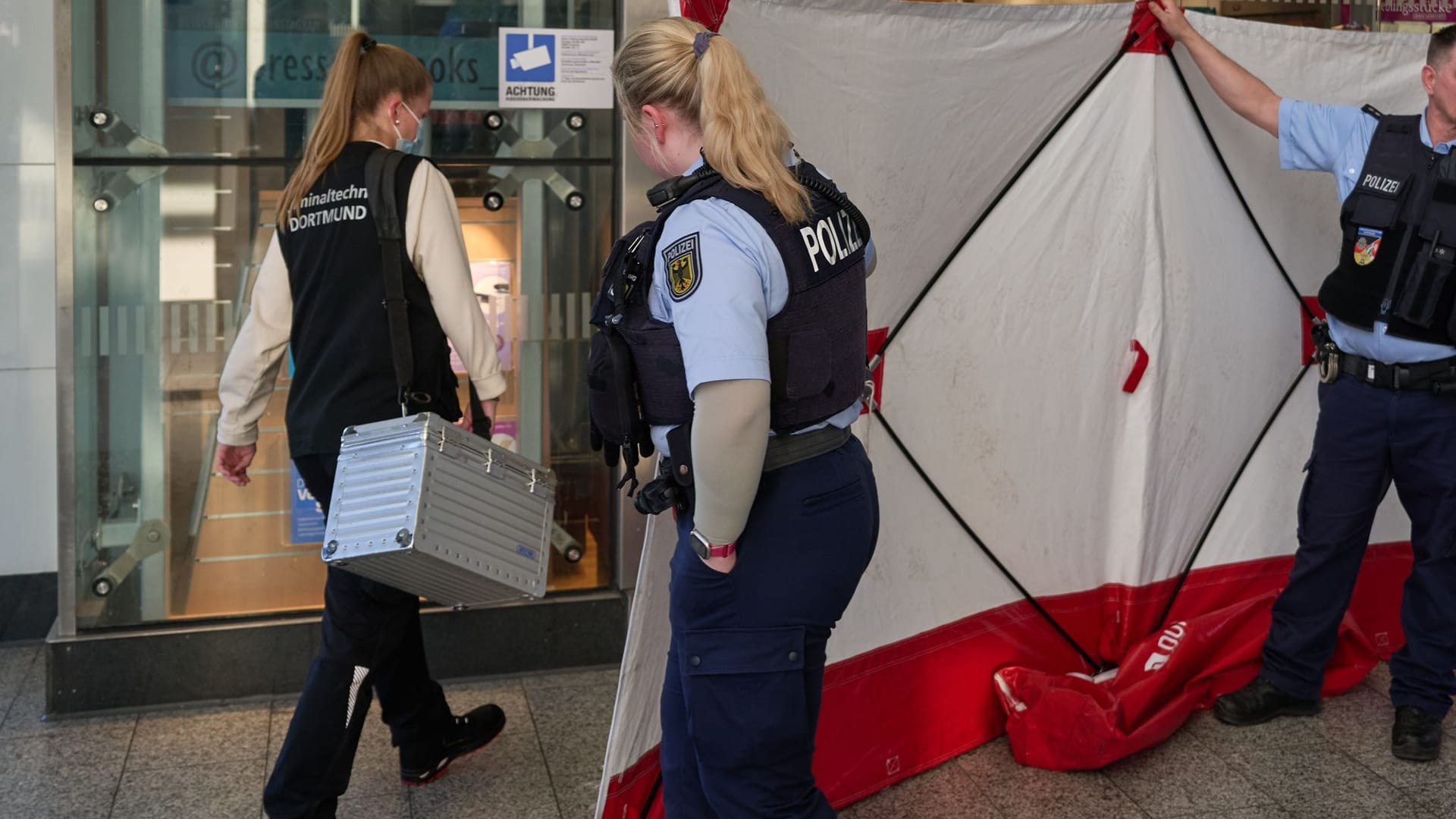 06.03.2025, Nordrhein-Westfalen, Dortmund: Polizisten öffnen den Sichtschutz vor einem Geschäft im Hauptbahnhof für eine Mitarbeiterin der Kriminaltechnik. Im Hauptbahnhof Dortmund ist am Mittag ein Mitarbeiter eines Fahrkartenschalters von einem Mann mit einem Messer angegriffen worden. Foto: Bernd Thissen/dpa +++ dpa-Bildfunk +++