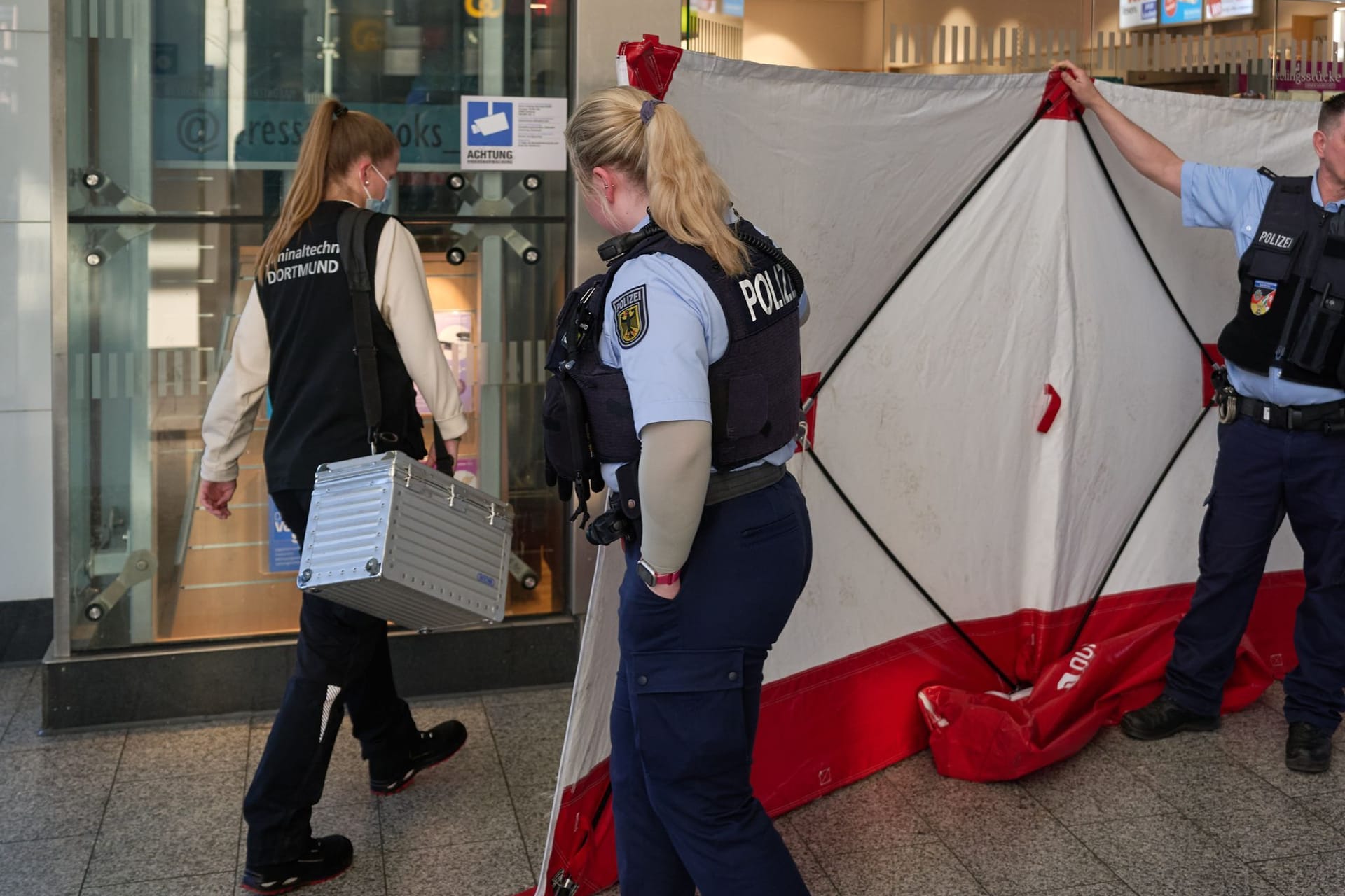 06.03.2025, Nordrhein-Westfalen, Dortmund: Polizisten öffnen den Sichtschutz vor einem Geschäft im Hauptbahnhof für eine Mitarbeiterin der Kriminaltechnik. Im Hauptbahnhof Dortmund ist am Mittag ein Mitarbeiter eines Fahrkartenschalters von einem Mann mit einem Messer angegriffen worden. Foto: Bernd Thissen/dpa +++ dpa-Bildfunk +++