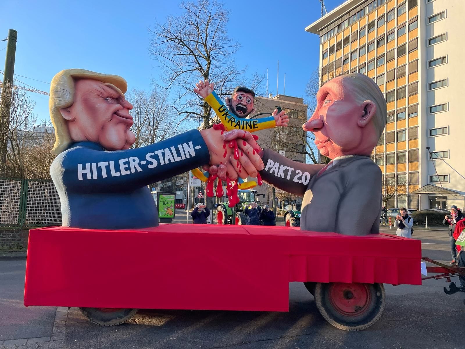 Ein Mottowagen zum Konflikt zwischen der Ukraine, Russland und den USA. Zu sehen sind Donald Trump, Präsident der Vereinigten Staaten, und Wladimir Putin, Präsident Russlands, beim Händeschütteln. Zwischen ihren Händen befindet sich eine Figur, mit der Aufschrift "Ukraine", die Präsident Selenskyj nachempfunden sein könnte. Sie ist in den Landesfarben Blau und Gelb gekleidet. Die Figur schreit und blutet, das Blut tropft die Hände der beiden Staatsmänner hinunter. Auf den Armen von Trump und Putin ist zudem zu lesen: "Hitler-Stalin-Pakt 2.0".