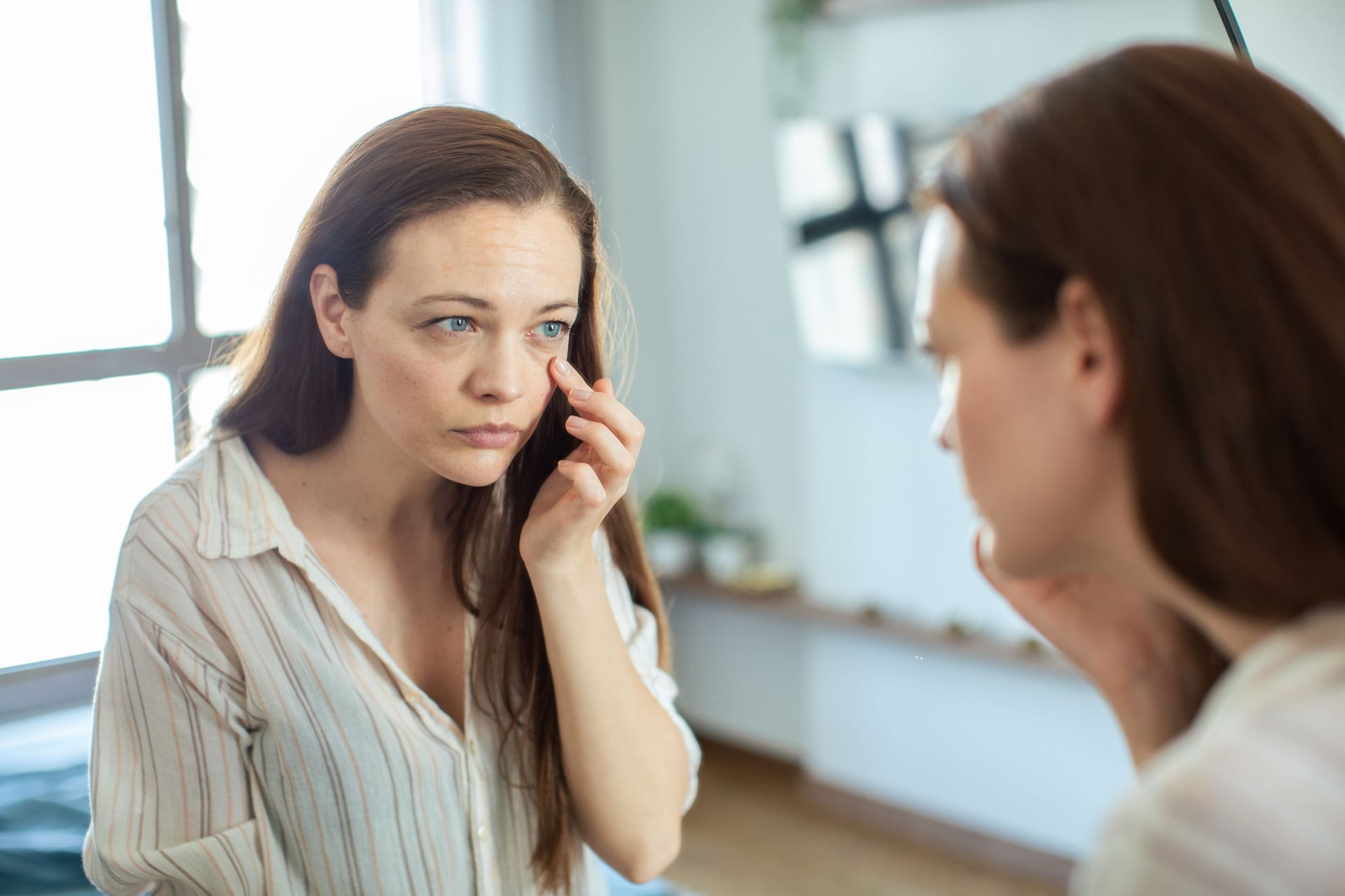 Eine Frau inspiziert ihr Auge im Spiegel: Für glasige, tränende Augen kann es viele Gründe geben.
