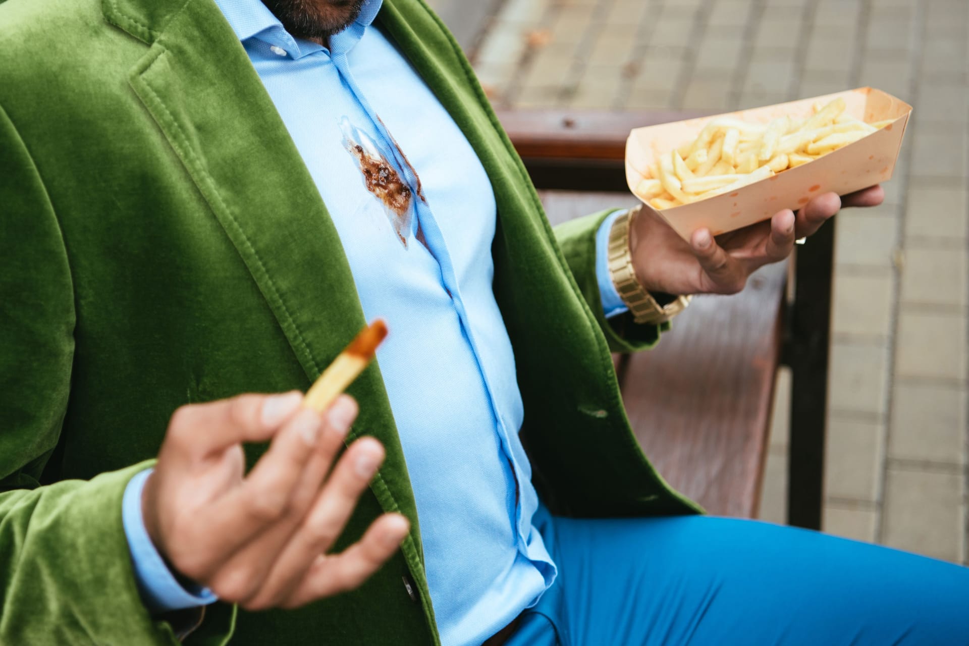 partial view of man in green velvet jacket with ketchup on shirt and french fries in hands sitting on bench on street