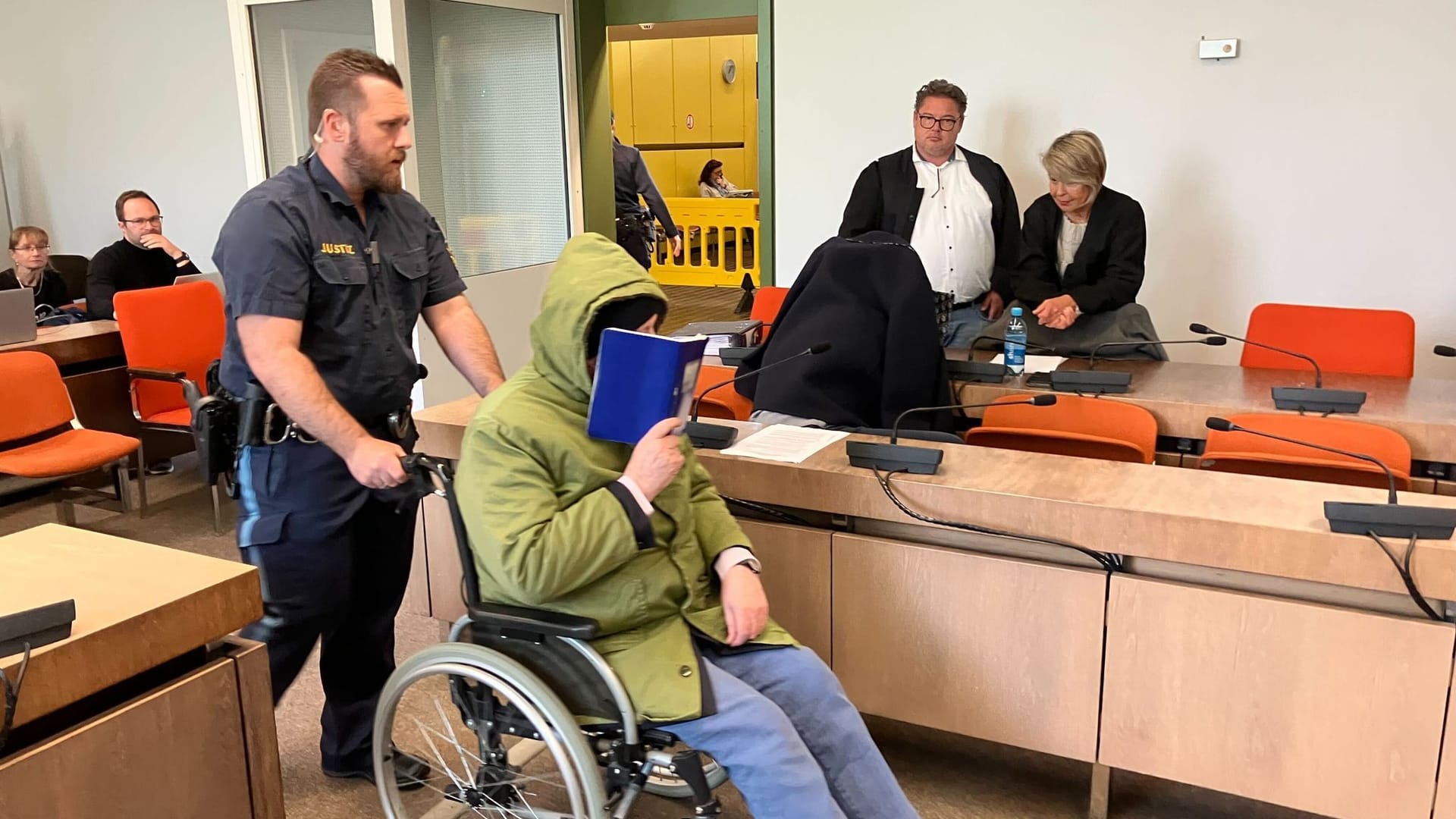 Ein Angeklagter wird auf einem Rollstuhl zur Urteilsverkündung im Prozess um Missbrauch von Kindern und Zwangsprostitution in den Saal des Landgerichts München I geschoben und verbirgt sein Gesicht. Ein weiterer Angeklagter versteckt sich auf der Anklagebank unter einer dunklen Jacke.