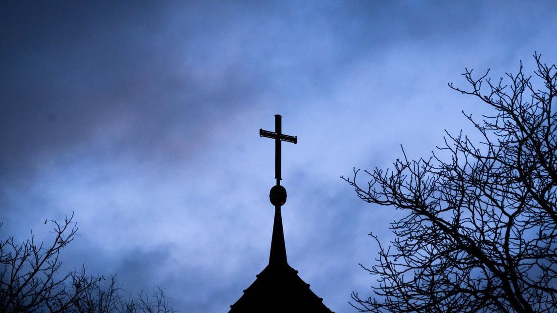 Ein Kreuz auf einer evangelischen Kirche, dahinter dunkle Wolken (Archivbild): Ein aktueller Bericht bringt neue Erkenntnisse im Fall des früheren Pastors Klaus Vollmer ans Licht.
