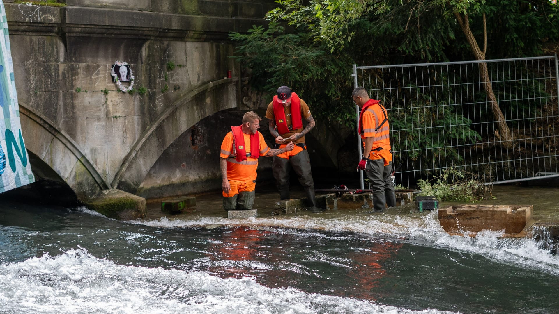 Mitarbeiter in Warnwesten und Watthosen bei Arbeiten an der Eisbachwelle: Die Öffnung unter der Brücke soll wieder geschlossen werden, um den Wasserfluss zu stabilisieren.