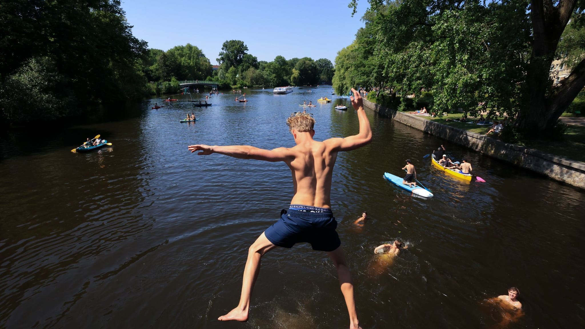 Ein junger Mann springt von einer Brücke in die Alster. Zahlreiche Menschen sind bei sommerlichen Temperaturen an und auf der Alster unterwegs.