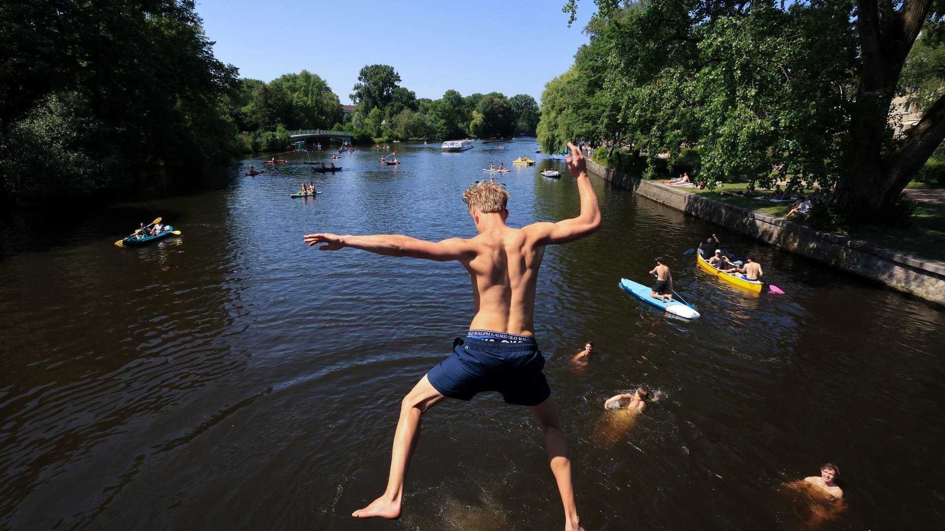 Ein junger Mann springt von einer Brücke in die Alster. Zahlreiche Menschen sind bei sommerlichen Temperaturen an und auf der Alster unterwegs.