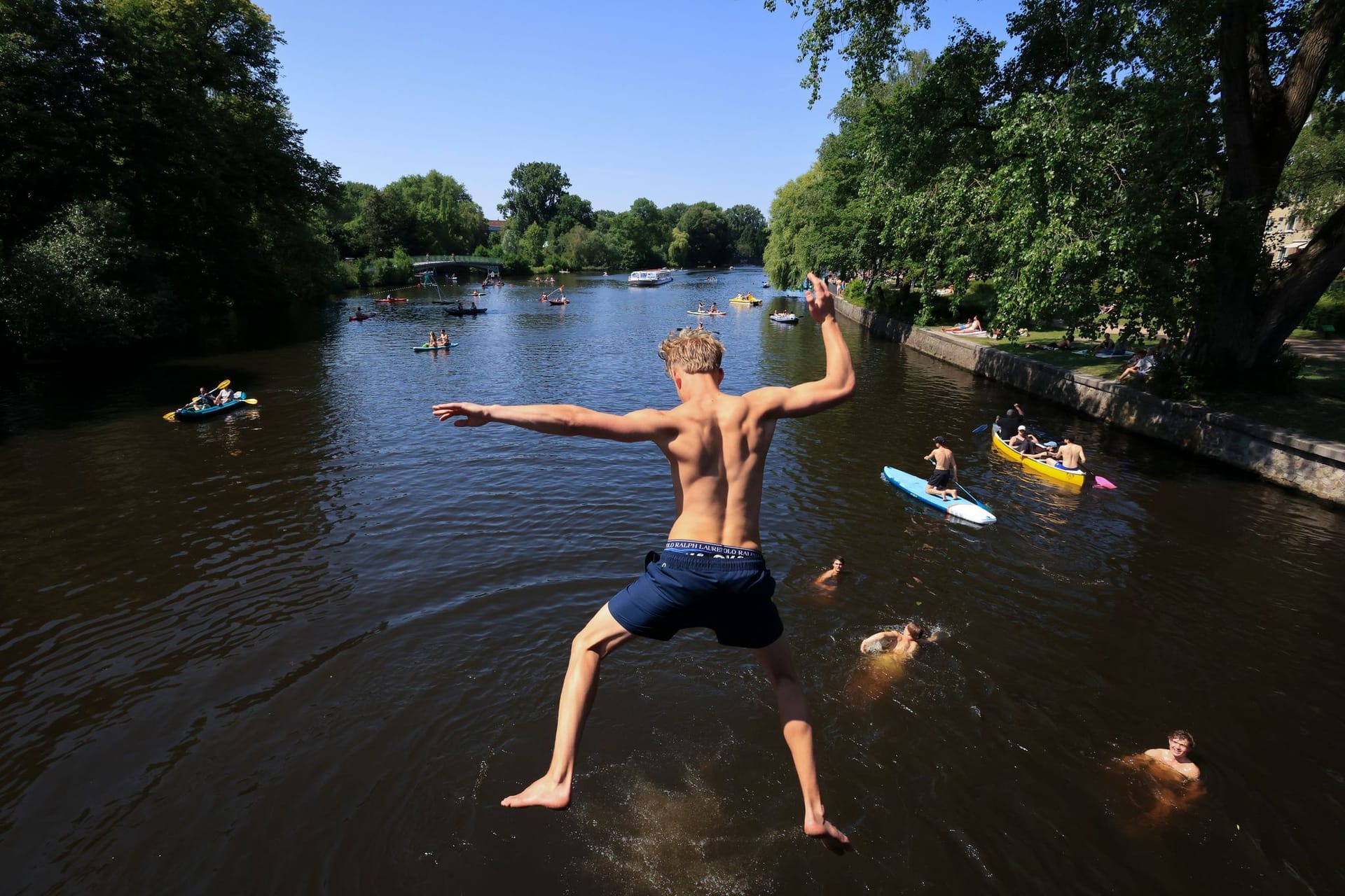 Ein junger Mann springt von einer Brücke in die Alster. Zahlreiche Menschen sind bei sommerlichen Temperaturen an und auf der Alster unterwegs.