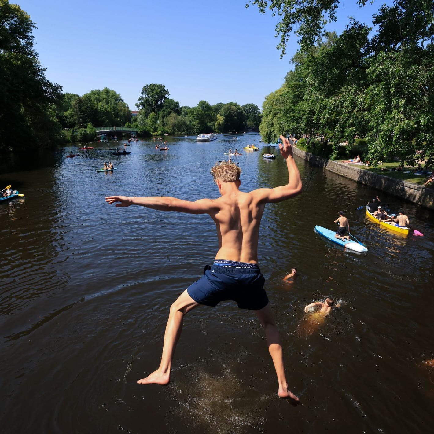 Ein junger Mann springt von einer Brücke in die Alster. Zahlreiche Menschen sind bei sommerlichen Temperaturen an und auf der Alster unterwegs.