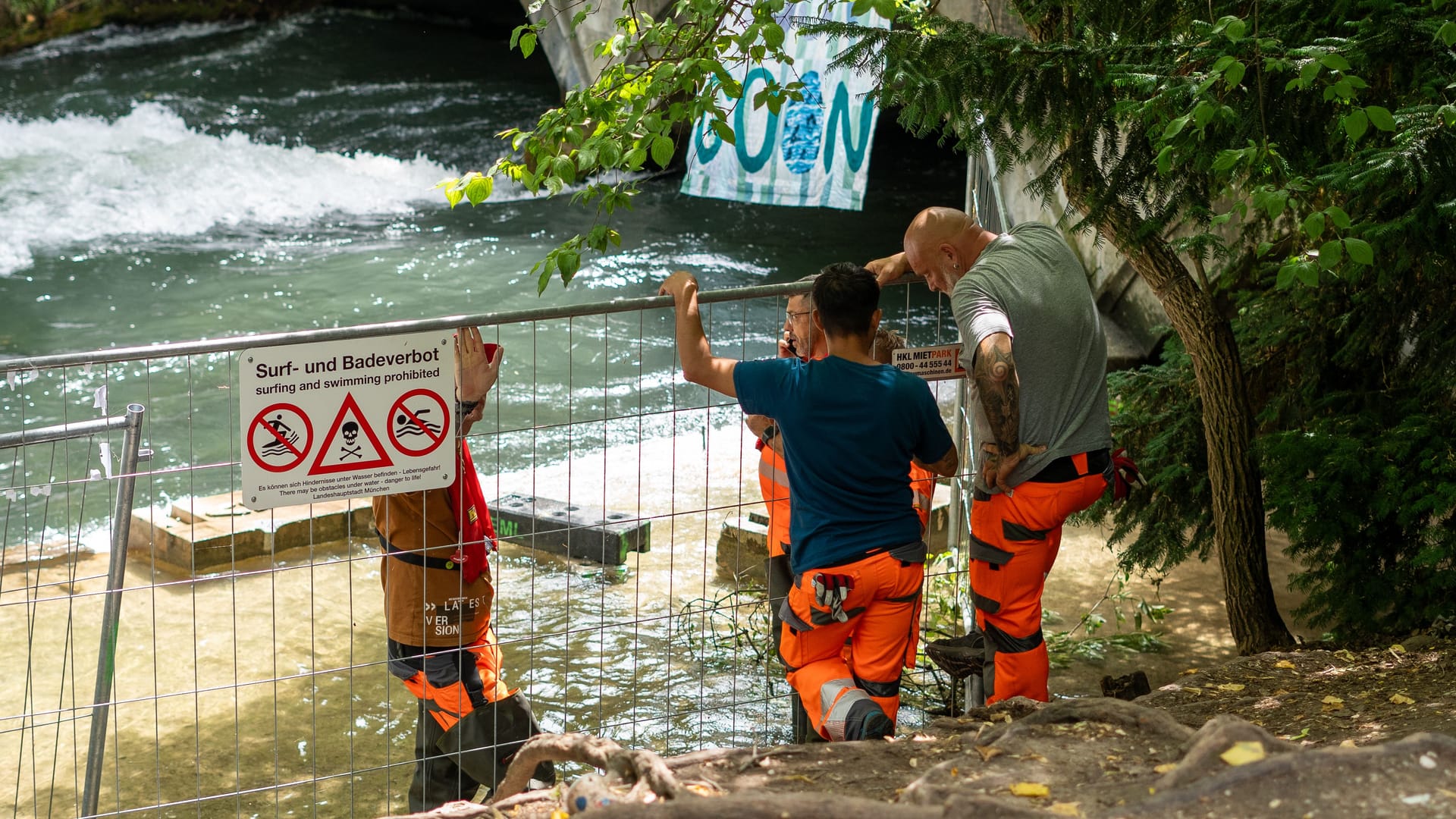 Absperrgitter mit Warnschild und Bauarbeiter am Eisbach: Trotz offizieller Freigabe bleibt die Welle gesperrt – Probleme verzögern den Neustart. Absperrgitter mit Warnschild und Bauarbeiter am Eisbach: Trotz offizieller Freigabe bleibt die Welle gesperrt – Probleme verzögern den Neustart.