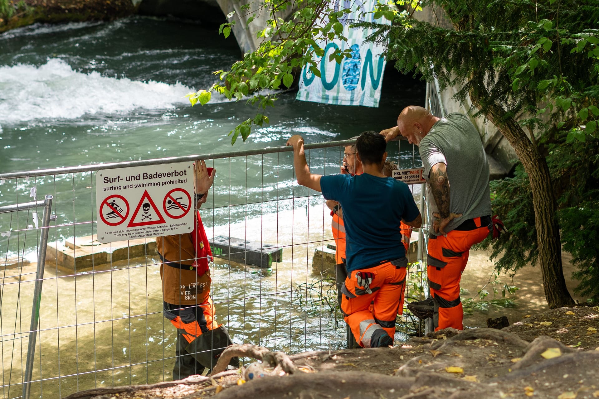 Absperrgitter mit Warnschild und Bauarbeiter am Eisbach: Trotz offizieller Freigabe bleibt die Welle gesperrt – Probleme verzögern den Neustart.