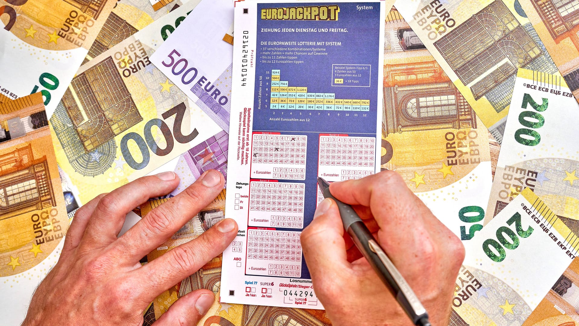 Mann füllt Eurojackpotspielschein vor einem Haufen Geldscheine aus (Symbolfoto): In Niedersachsen haben zwei Spieler jeweils große Summen gewonnen.