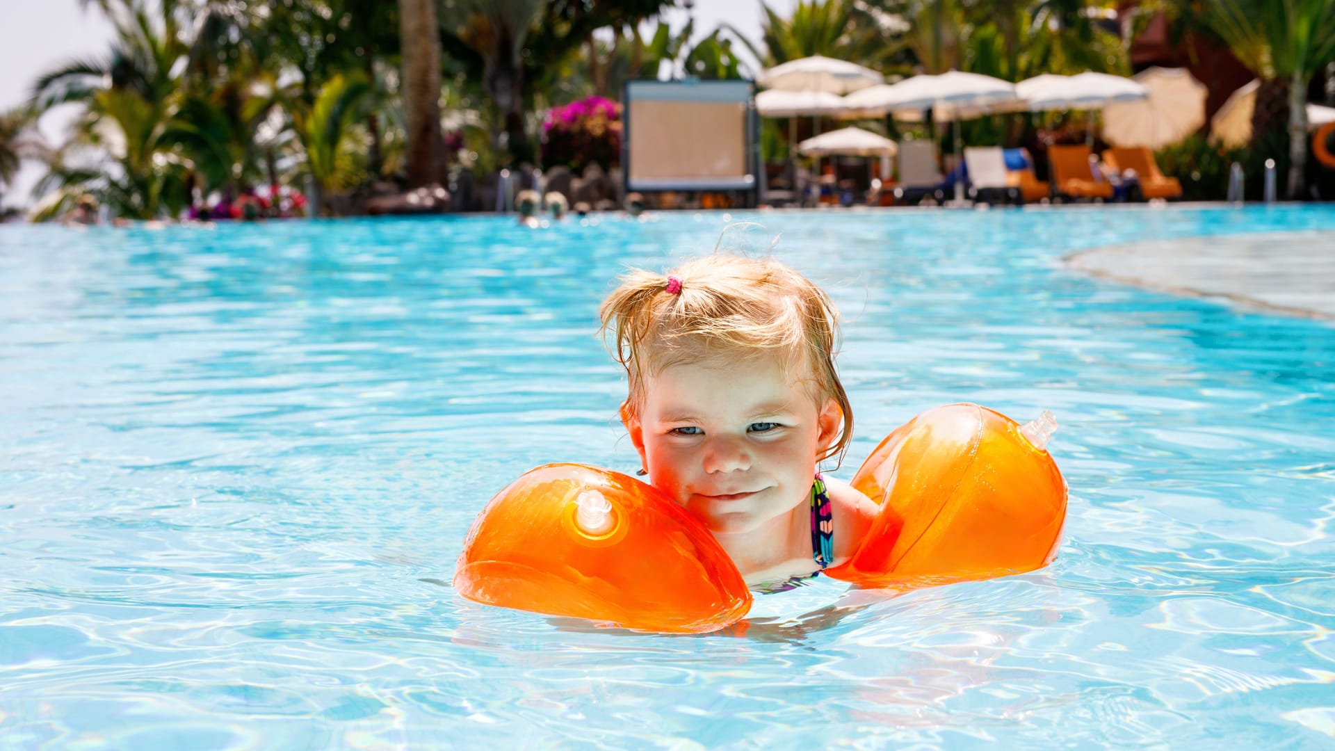 Little toddler girl with protective swimmies playing in outdoor swimming pool by sunset. Baby Child learning to swim in outdoor pool, splashing with water, laughing and having fun. Family vacations. Little toddler girl with protective swimmies playing in outdoor swimming pool by sunset. Baby Child learning to swim in outdoor pool, splashing with water, laughing and having fun. Family vacations.