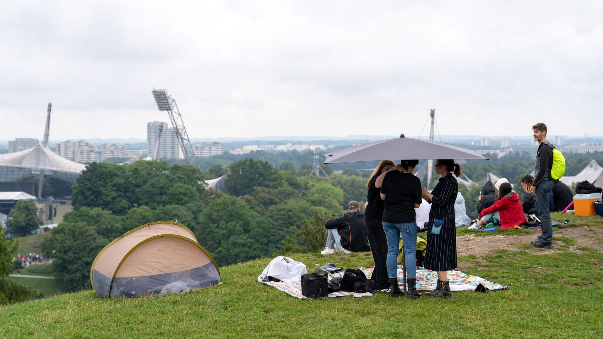 Eine Gruppe baut auf dem Olympiaberg einen großen Schirm auf: Stunden vor dem Robbie-Williams-Konzert sichern sich Fans in München möglichst trockene Plätze mit Aussicht aufs Stadion.