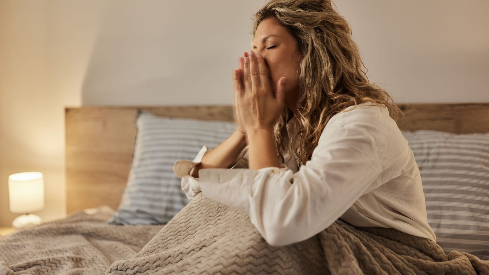 Eine Frau sitzt im Bett und bedeckt die Nase mit den Händen: Geht die Nase nachts ständig zu, fällt das Schlafen oft schwer. Eine Frau sitzt im Bett und bedeckt die Nase mit den Händen: Geht die Nase nachts ständig zu, fällt das Schlafen oft schwer.